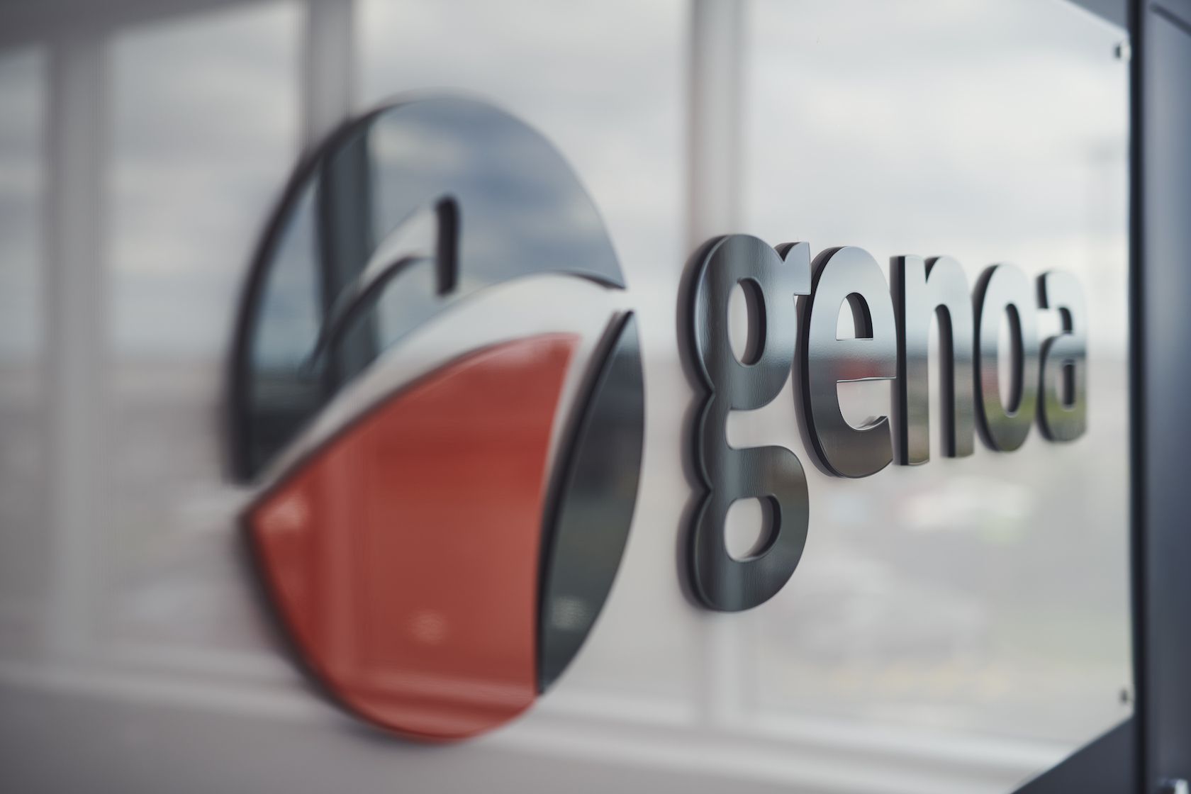 Close-up of a reflective Genoa logo on glass with blurred office background, emphasizing modern corporate branding and design.