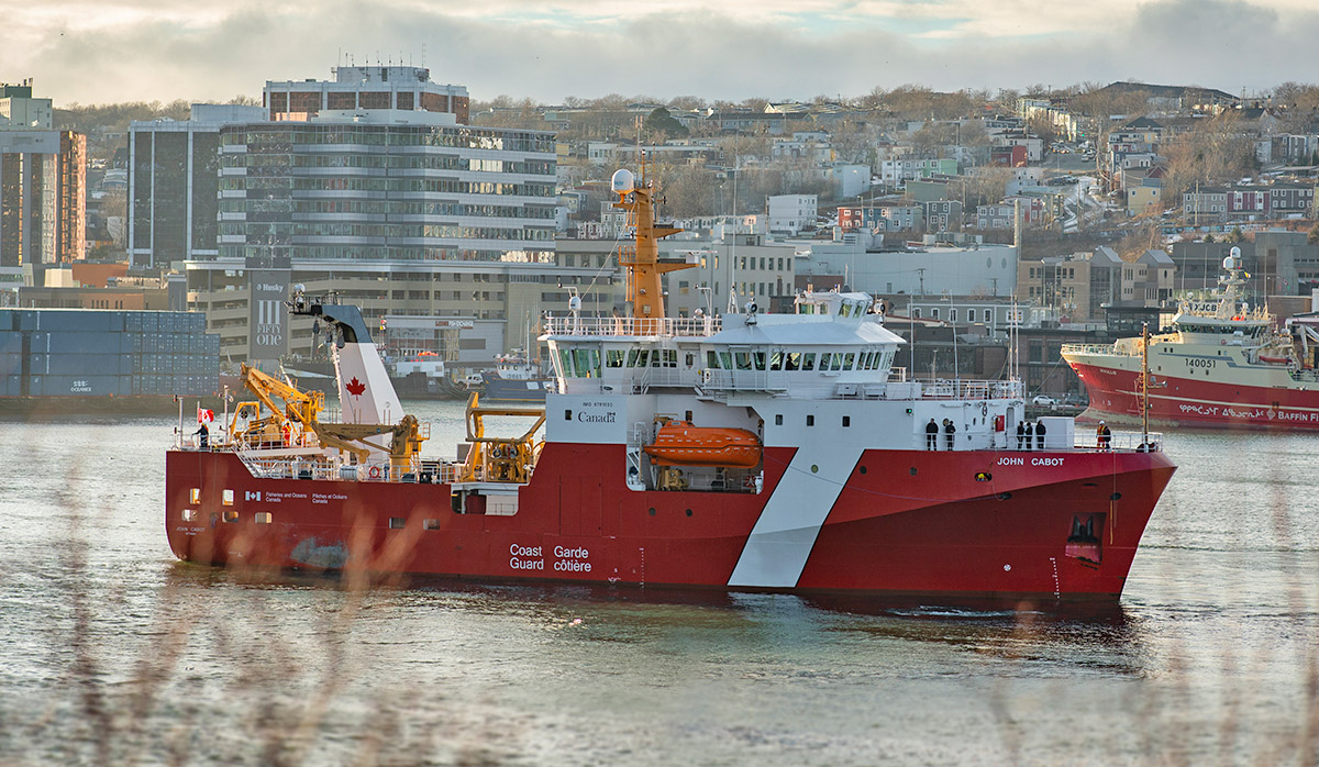 A large red and white Coast Guard ship named "John Cabot" in a harbor with a cityscape in the background.