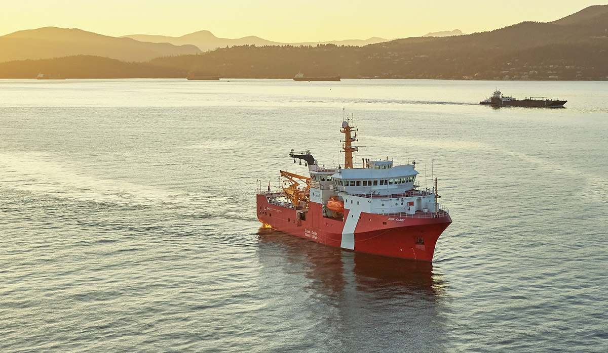 A red and white research vessel sailing on calm water at sunset with mountains in the background, another ship is visible in the distance.