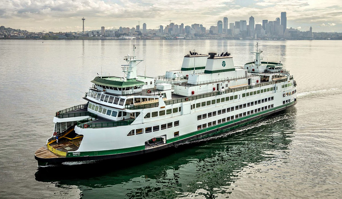 A large green and white ferry boat sailing on calm waters with the Seattle skyline, including the Space Needle, in the background.