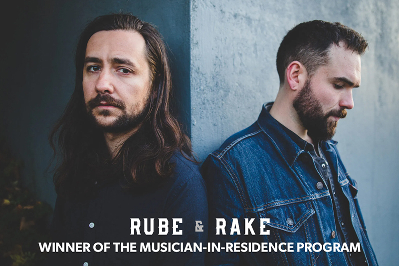 Two individuals standing against a blue wall, both dressed in dark clothing with one wearing a denim jacket. White text over the image reads "RUBE RAKE, WINNER OF THE MUSICIAN-IN-RESIDENCE PROGRAM."