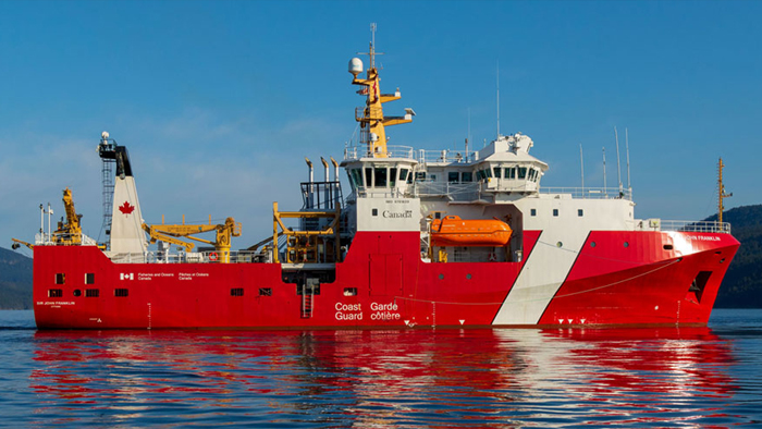 A red and white Canadian Coast Guard ship floating on calm blue water with a mountain range in the background.