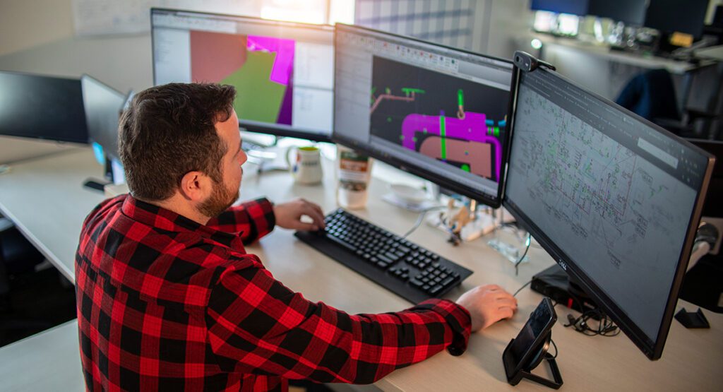 Male software developer in red plaid shirt working on dual monitors displaying code and graphics in a modern tech office environment.