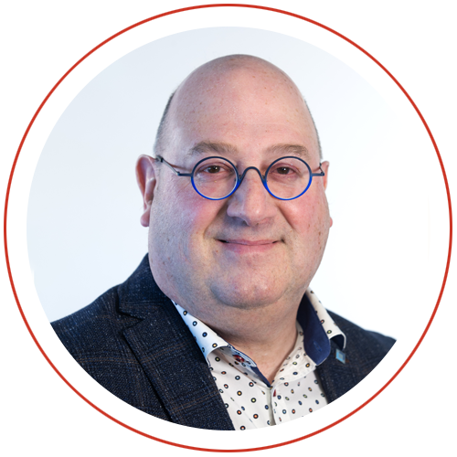 Professional portrait of a smiling man with glasses wearing a blazer and polka dot shirt, isolated on a white background.