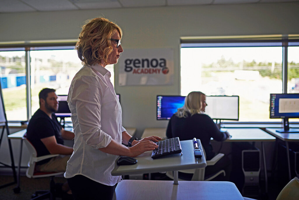 Focused professional woman using a computer at a standing desk in the modern Genoa Academy office, teaching or supervising in a tech educational environment.