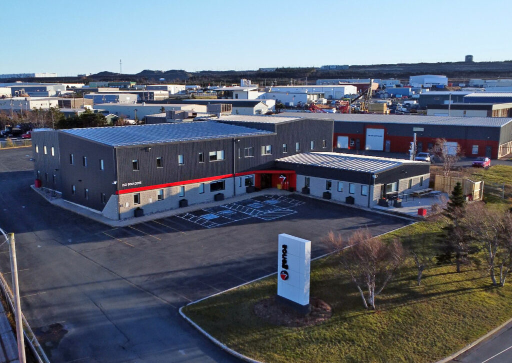 Aerial view of Dundee featuring a spacious parking lot and several business buildings, prominently displaying the Genoa Design International office with ISO 9001:2015 certification signage, with green landscape under a clear blue sky.