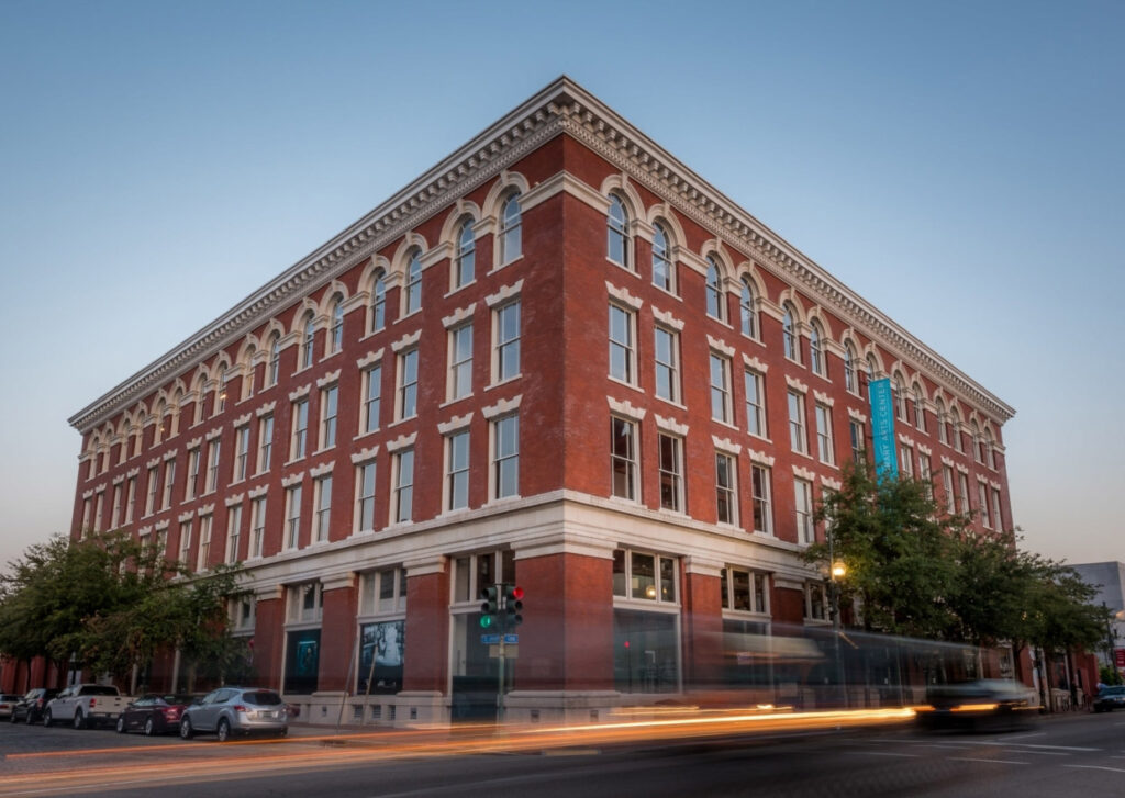 A red brick historic building on a street corner with arched windows and white trim, evening sky with long exposure of a bus passing by creating light trails.