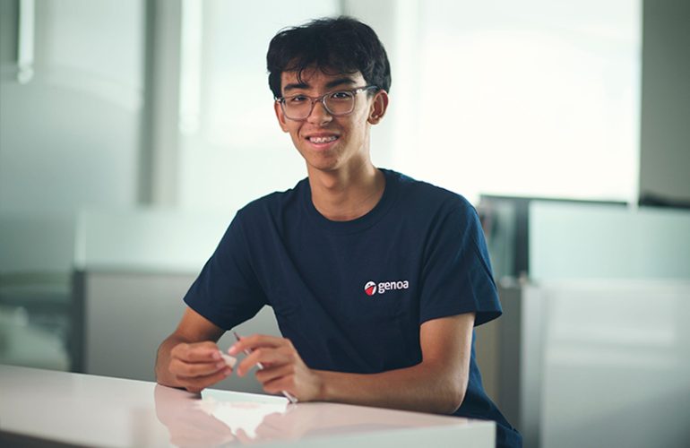 Smiling young male student with glasses wearing blue Genoa logo t-shirt sitting at a white table in a bright modern room.