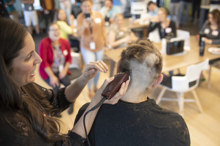 A person getting their hair cut with clippers in an indoor setting surrounded by people sitting and standing with mugs and plates in their hands.