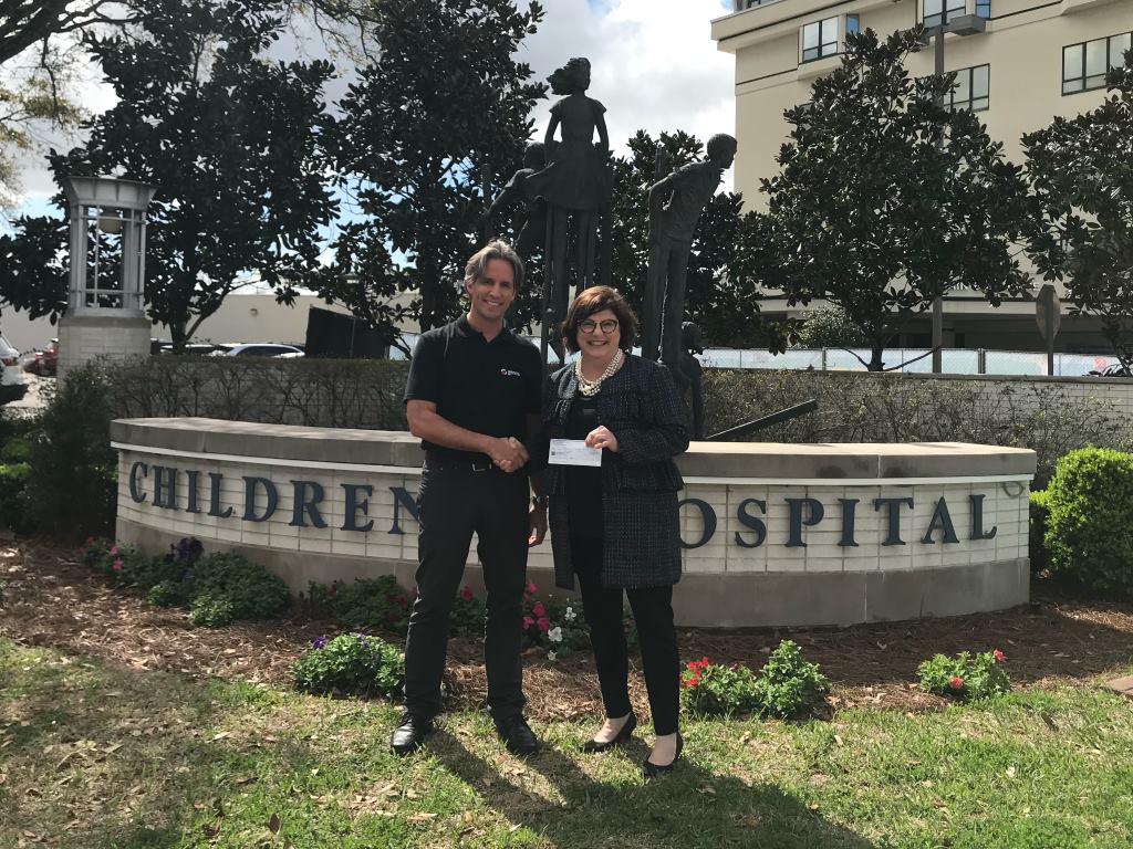 A man and a woman stand in front of a semi-circular stone sign with the inscription "CHILDREN'S HOSPITAL," behind them is a sculpture of children, and the scene is set against a backdrop of trees, shrubbery, and a multi-story building under a clear sky.