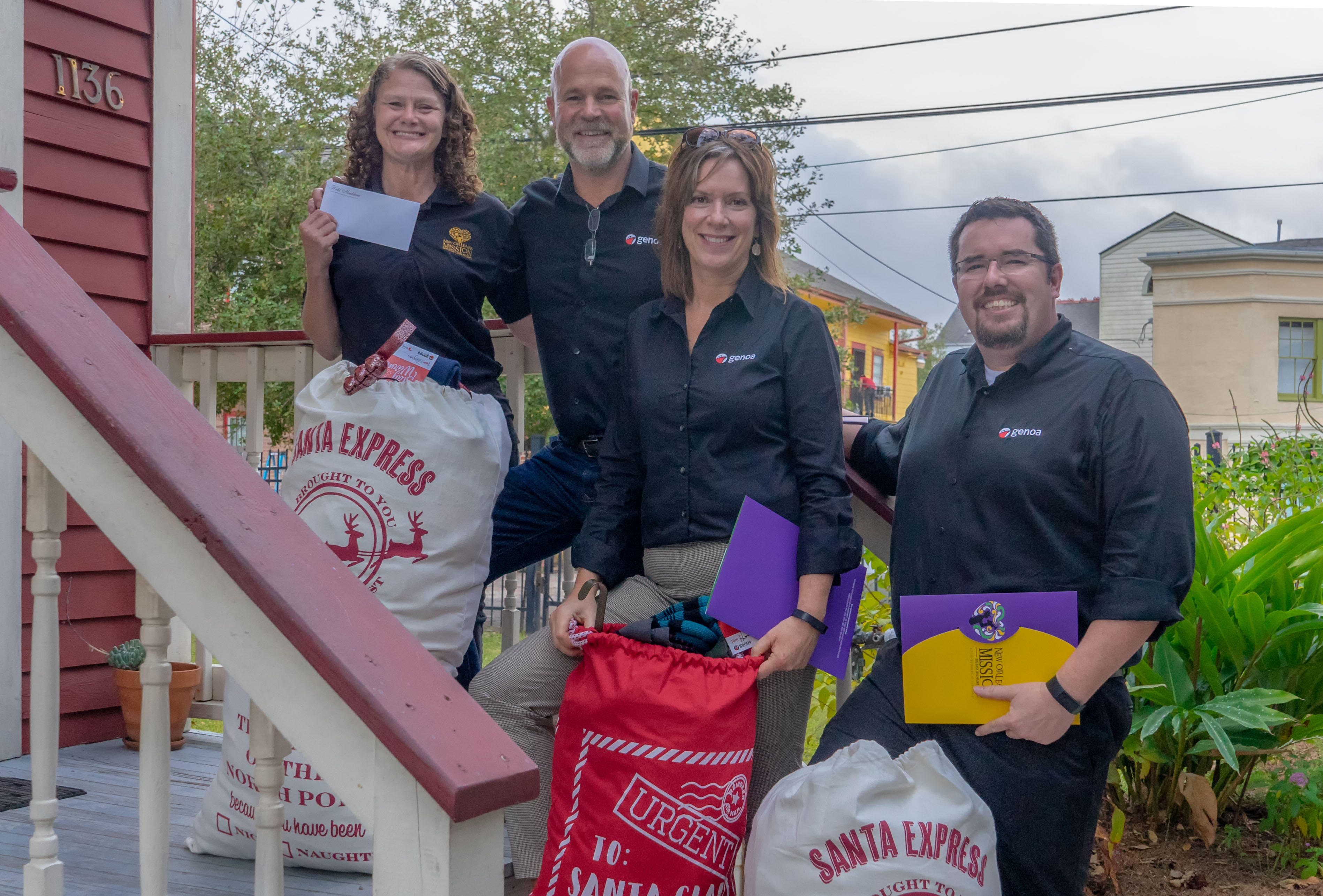 Four smiling people in company shirts holding gift bags and an envelope on a residential porch with festive Santa Express bags.