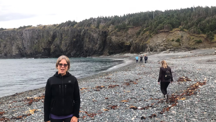 Five people on a rocky beach, one women dressed in black with sunglasses facing towards the camera in the forefront.