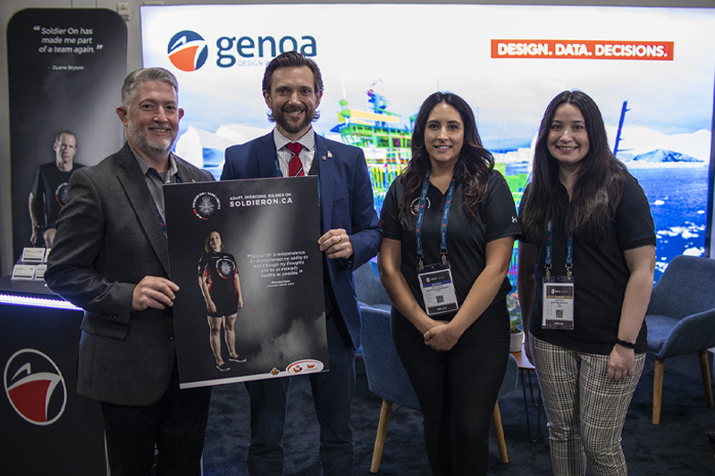 Four professionals, two men and two women, stand in a conference booth for Genoa Design featuring a poster for Soldier On initiative.