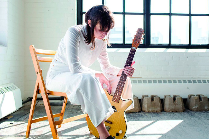 A person in a white dress sitting on a wooden chair while holding a guitar in a room with large windows and bright natural light.