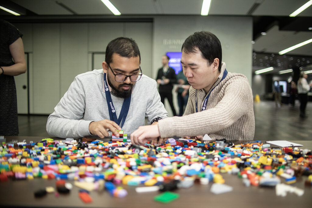 Two men at a table building with Lego pieces in a big room.