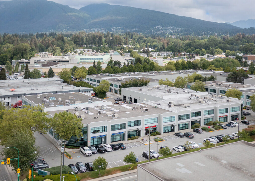 Aerial view of modern industrial building with spacious parking lot near forested mountain range, showcasing flat buildings with numerous air conditioning units on roofs, under overcast sky.