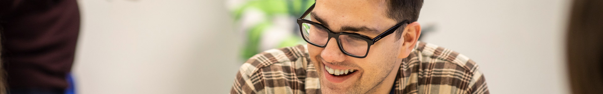 A man wearing a brown and beige plaid shirt and glasses, sitting in front of a blurred background.