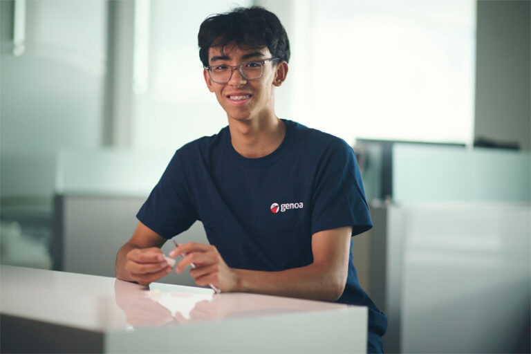 Smiling young male student with glasses wearing blue Genoa logo t-shirt sitting at a white table in a bright modern room.