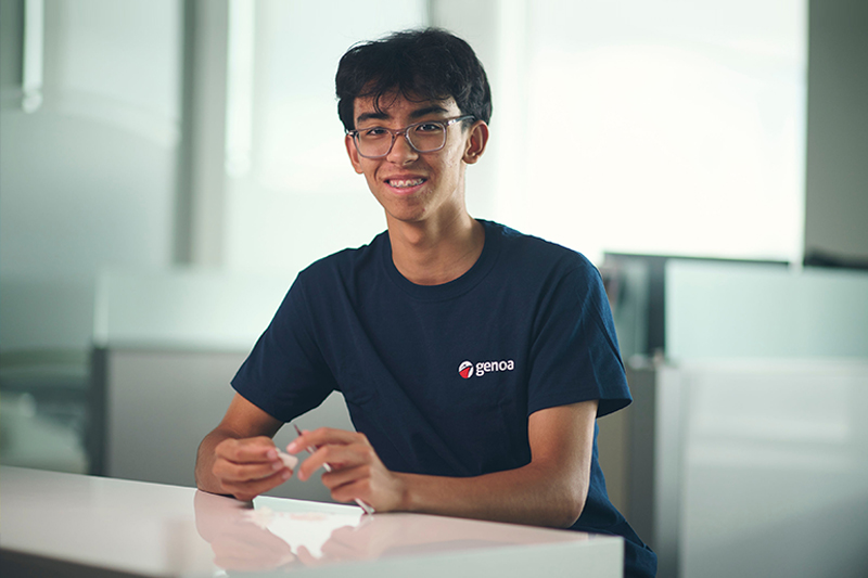 Smiling young male student with glasses wearing blue Genoa logo t-shirt sitting at a white table in a bright modern room.