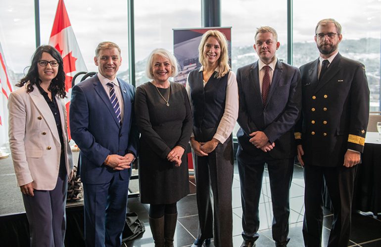 Group of professionals standing in a row indoors with a backdrop featuring national flags and a scenic view through a window behind them.