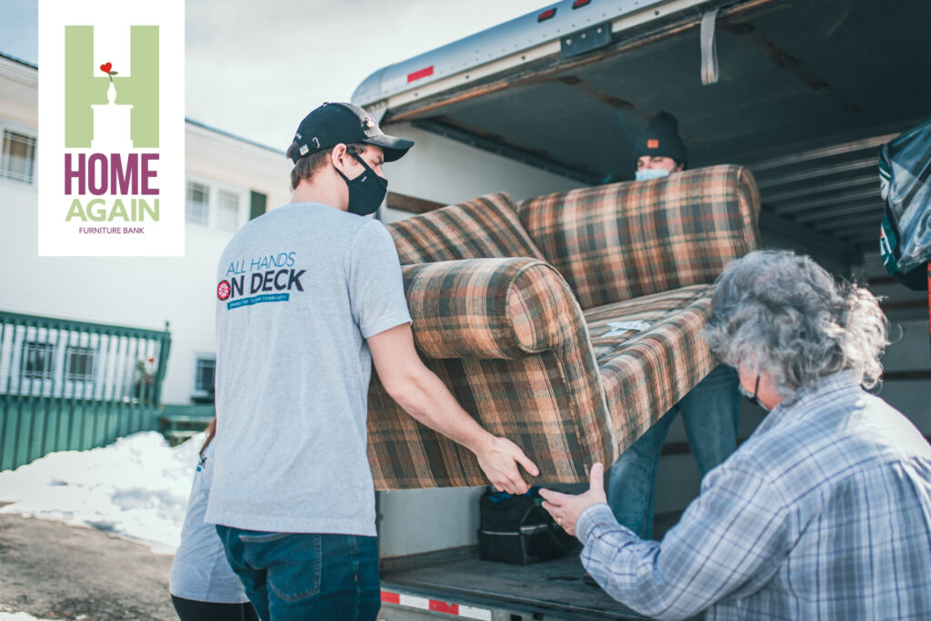 Two men carrying a plaid couch into a moving truck, wearing face masks, with the Home Again Furniture Bank logo in the corner in support of Genoa's community support initatives.