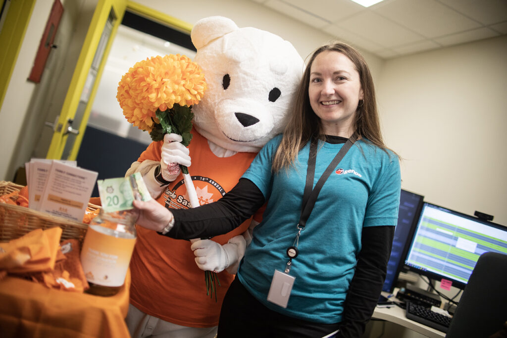 A person in a bear mascot costume holding a bouquet of flowers standing next to a women advocating for community support, wearing a blue long-sleeved shirt with a lanyard ID, computer screen in the background, next to the bear there is a table with an orange table cloth and a basket on top.