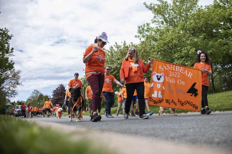 A group of individuals wearing bright orange shirts participating in the "Sashbear Walk On The Rock 2022. Together We Are Stronger" event, with some walking dogs on leashes and others holding a large orange banner featuring a cartoon bear.