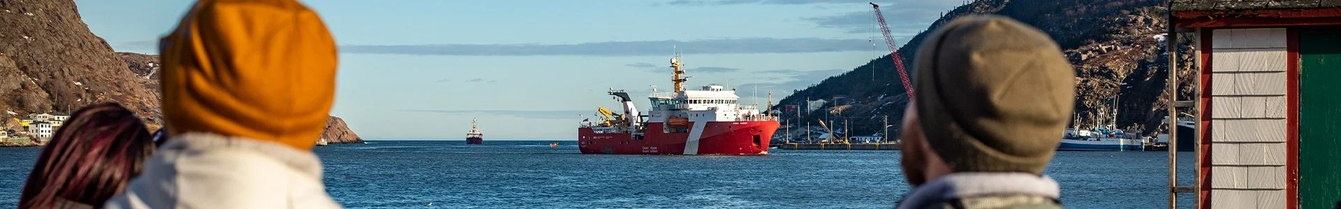 Two people wearing beanies looking at a red and white ship on the water in a harbor with mountains in the background.