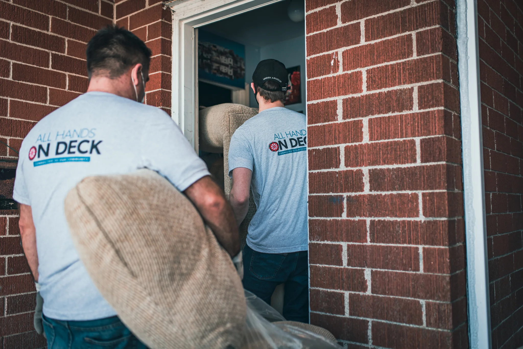 Two movers carrying a sofa into a house through a doorway, featuring All Hands on Deck t-shirts.